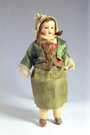 Κούκλα με κεφάλι από πορσελάνη “bisque” και αρθρωτά μέλη από μίγμα “composition”, γερμανικού εργοστασίου, ντυμένη με την τοπική γυναικεία φορεσιά της Κιμώλου. Αποτελεί τμήμα της συλλογής με κούκλες, τις οποίες η βασίλισσα Όλγα δώρισε στο ΛτΕ το 1914. ΜΕΛΕ, A.M. 14493.